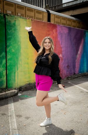 Young woman in pink shorts and black top poses joyfully in front of a colorful rainbow mural outdoors.