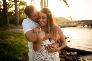 Couple embracing at golden hour sunset by the water, woman in lace dress smiling as man kisses her cheek.