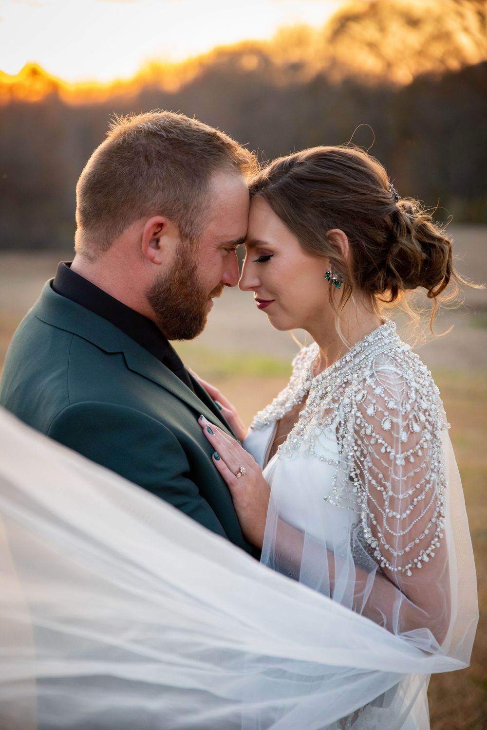 Bride and groom touching foreheads at golden sunset, bride in beaded cape veil wedding gown, groom in dark green suit.