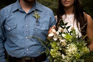 Couple at wedding, groom in blue chambray shirt with boutonniere, bride holding lush white and green floral bouquet.