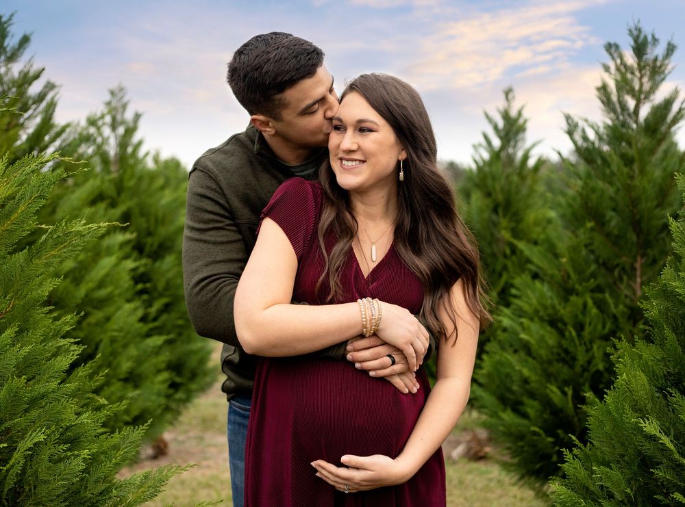 Couple embracing in a Christmas tree farm, pregnant woman in burgundy dress with partner kissing her cheek.