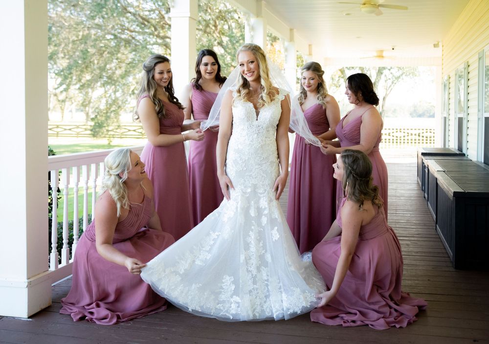 Bride in white lace mermaid gown surrounded by bridesmaids in dusty rose dresses on a classic white porch.