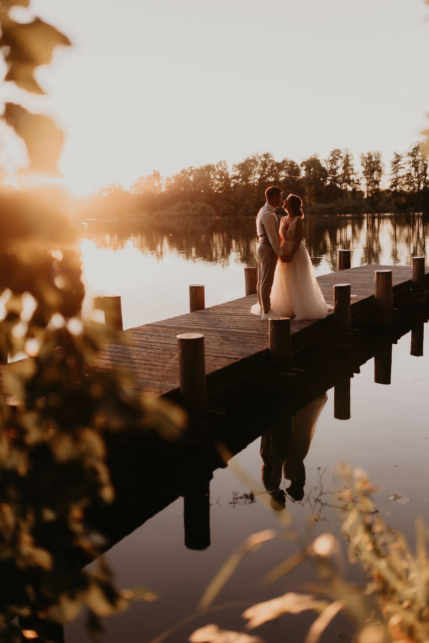 Couple sharing a romantic embrace on a wooden dock at golden sunset, reflected in calm lake waters surrounded by trees.