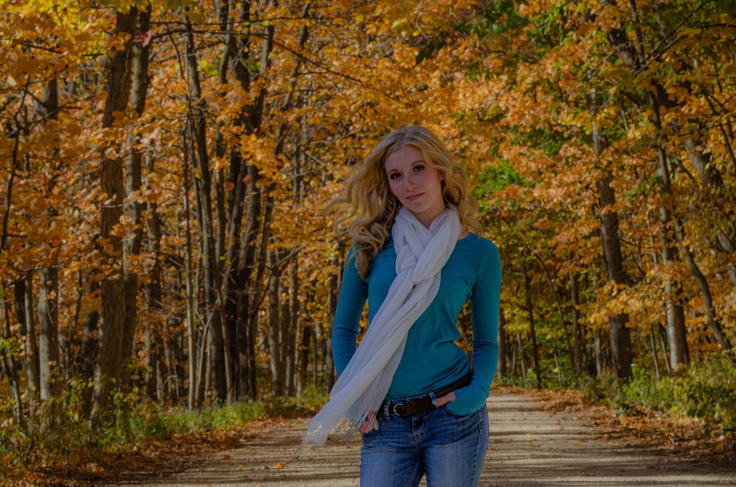 Blonde woman in teal sweater and white scarf standing on a tree-lined path surrounded by vibrant autumn foliage.