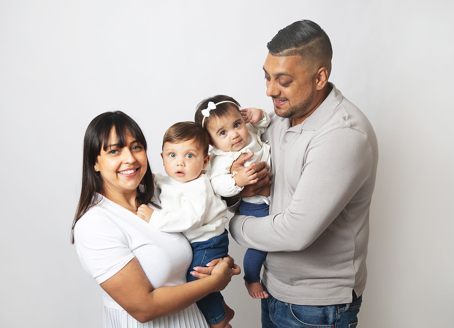 Family portrait of parents holding two young toddlers, smiling together against a white background. During a family photoshoot in Bexley