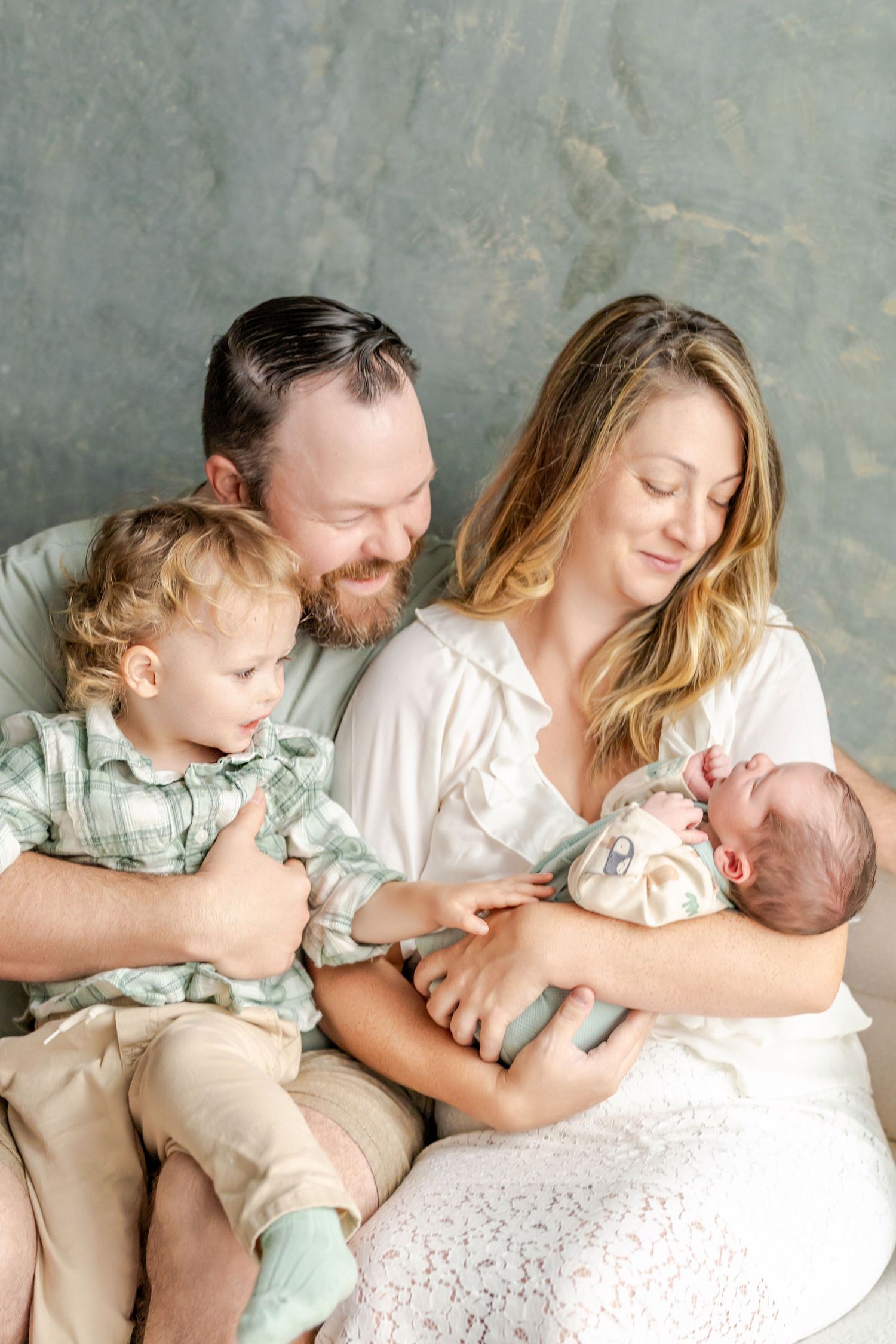 A happy family of four with a father, mother, toddler, and newborn baby posing together for a portrait.