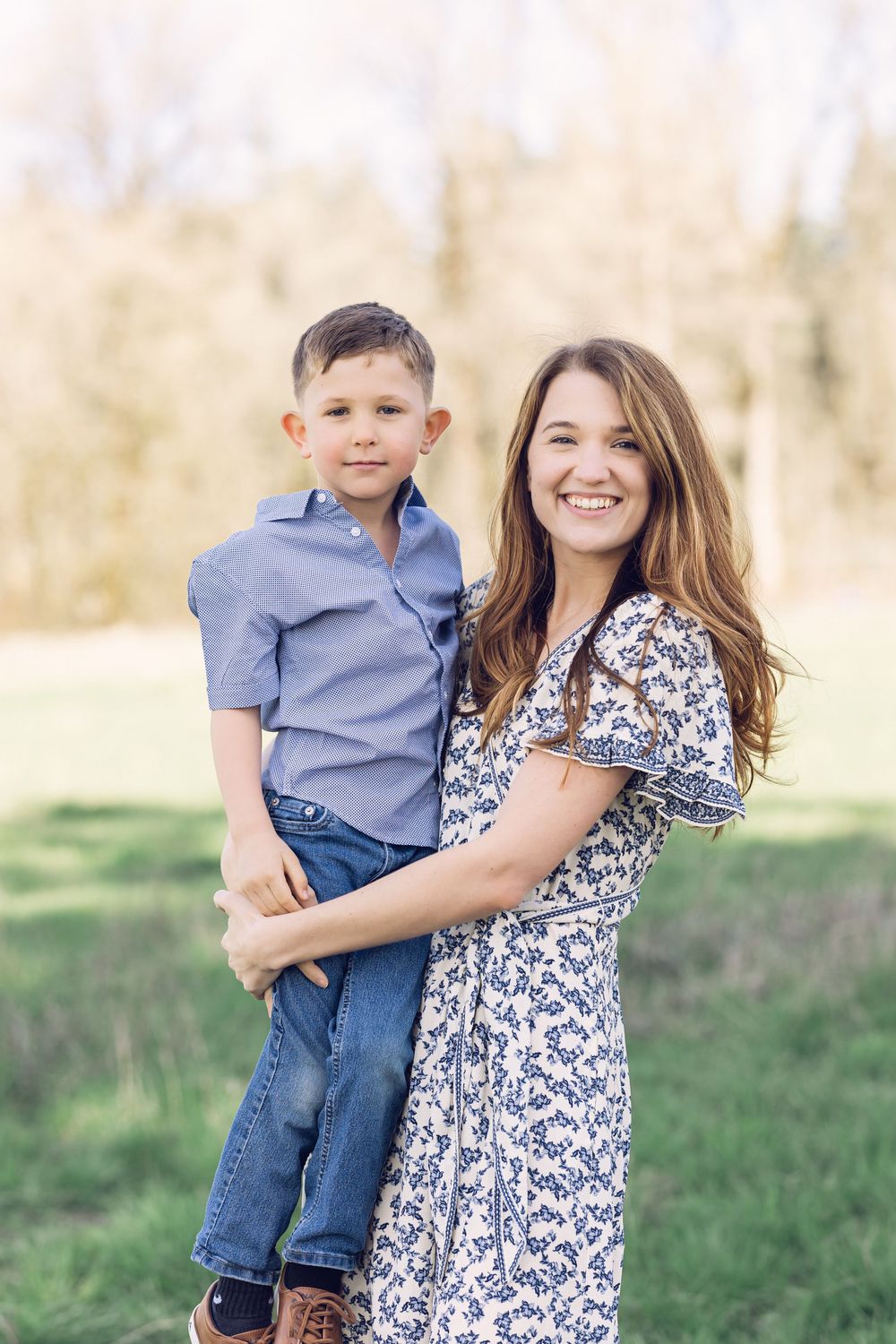 A smiling mother in a floral dress holds her young son in a blue shirt outdoors.