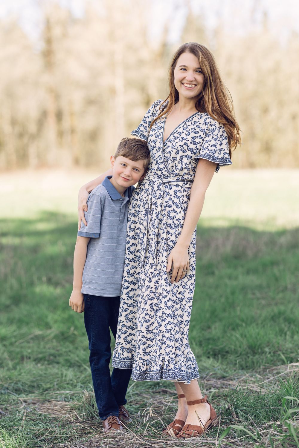 A happy mother in a floral dress stands with her son in a gray polo shirt in a park.