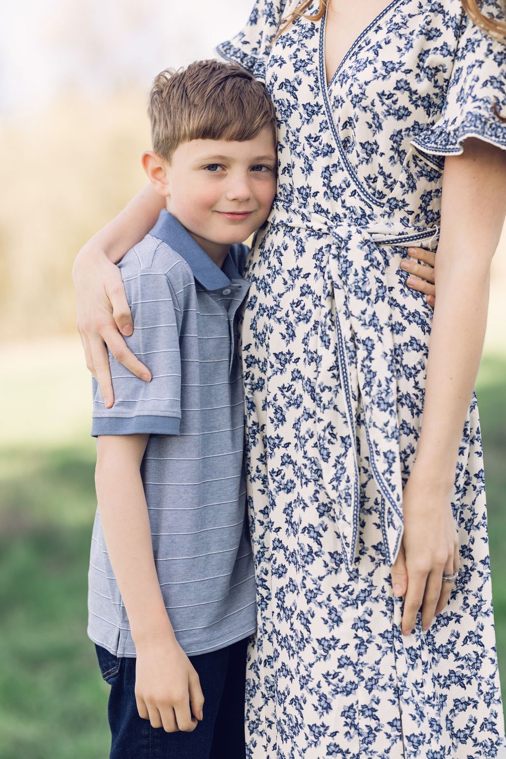 A young boy in a striped polo leans against his mothers floral dress in a sunny park.