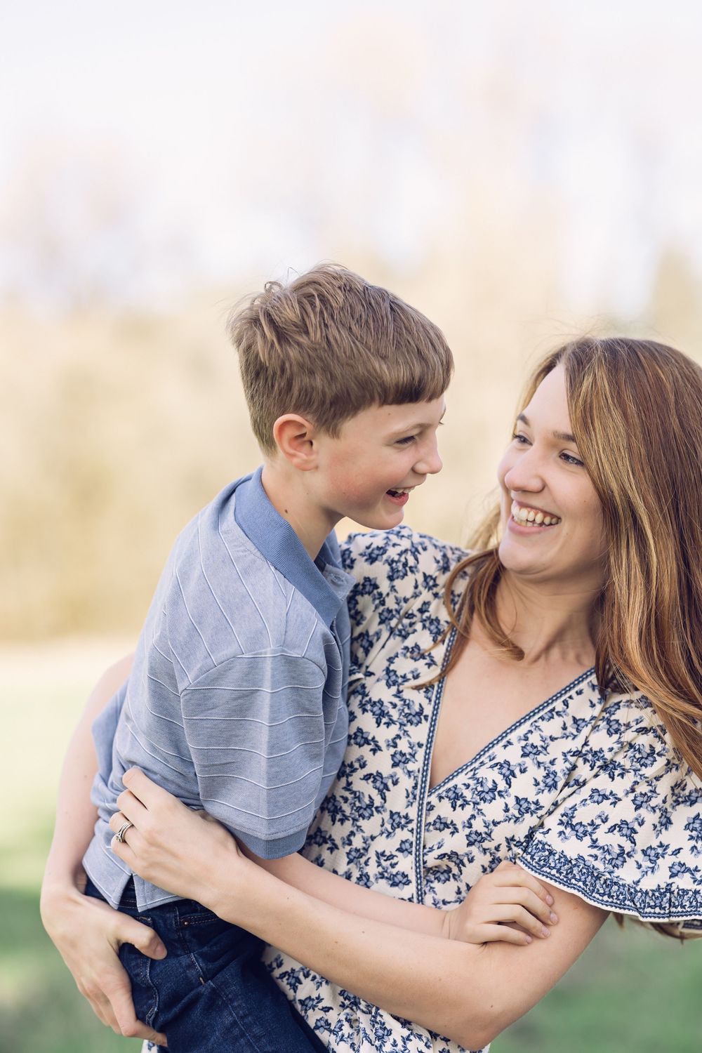 A mother and son laugh together while embracing in a sunlit outdoor setting.