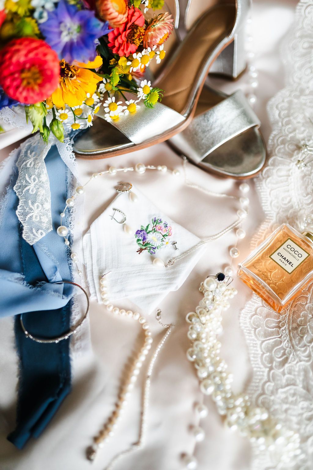Bridal flat lay with silver heels, colorful bouquet, pearl jewelry, blue ribbon, embroidered handkerchief, and Chanel Coco perfume.