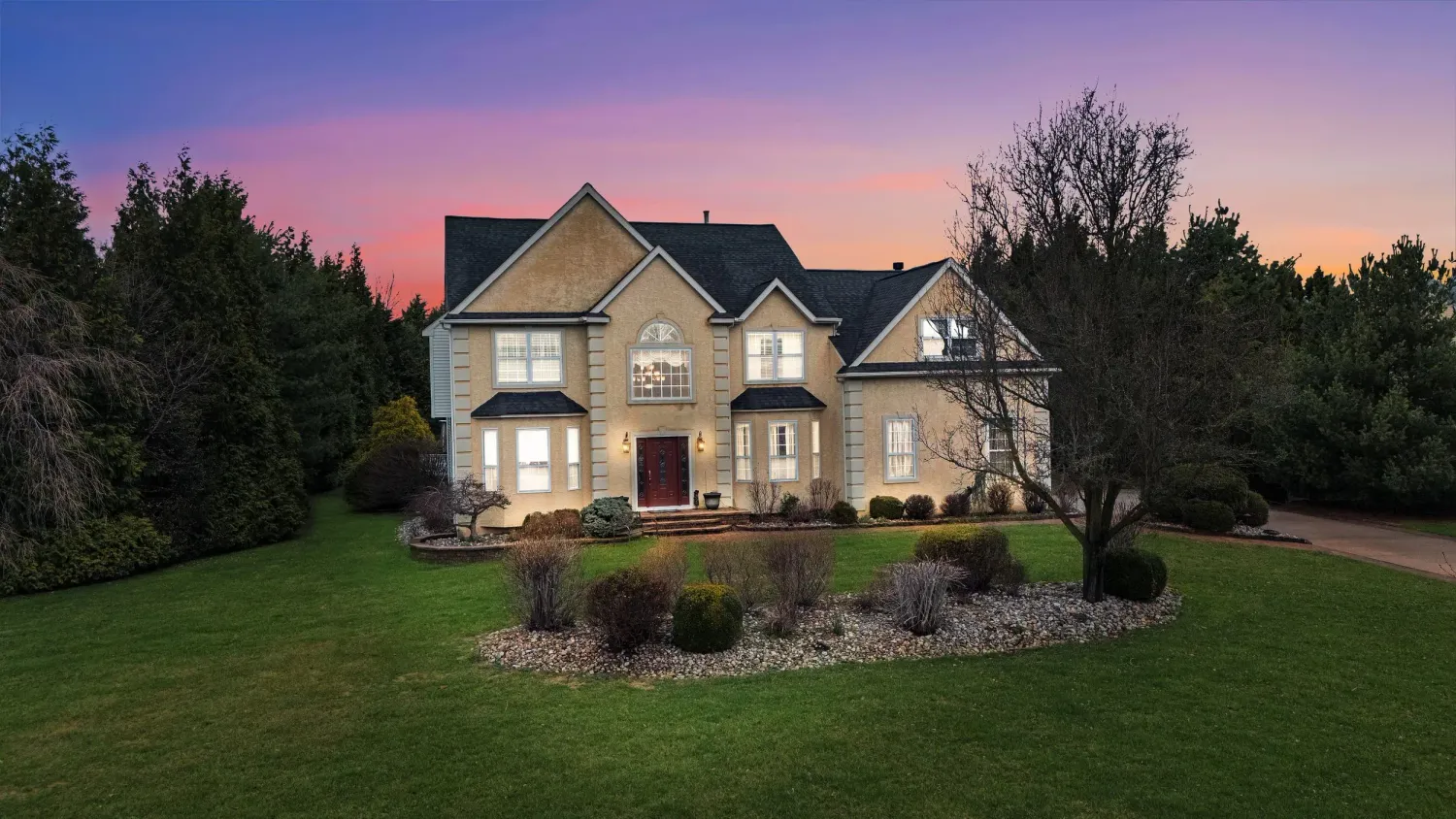 Large two-story stone home with manicured lawn at twilight, featuring warm interior lighting and lush landscaping.