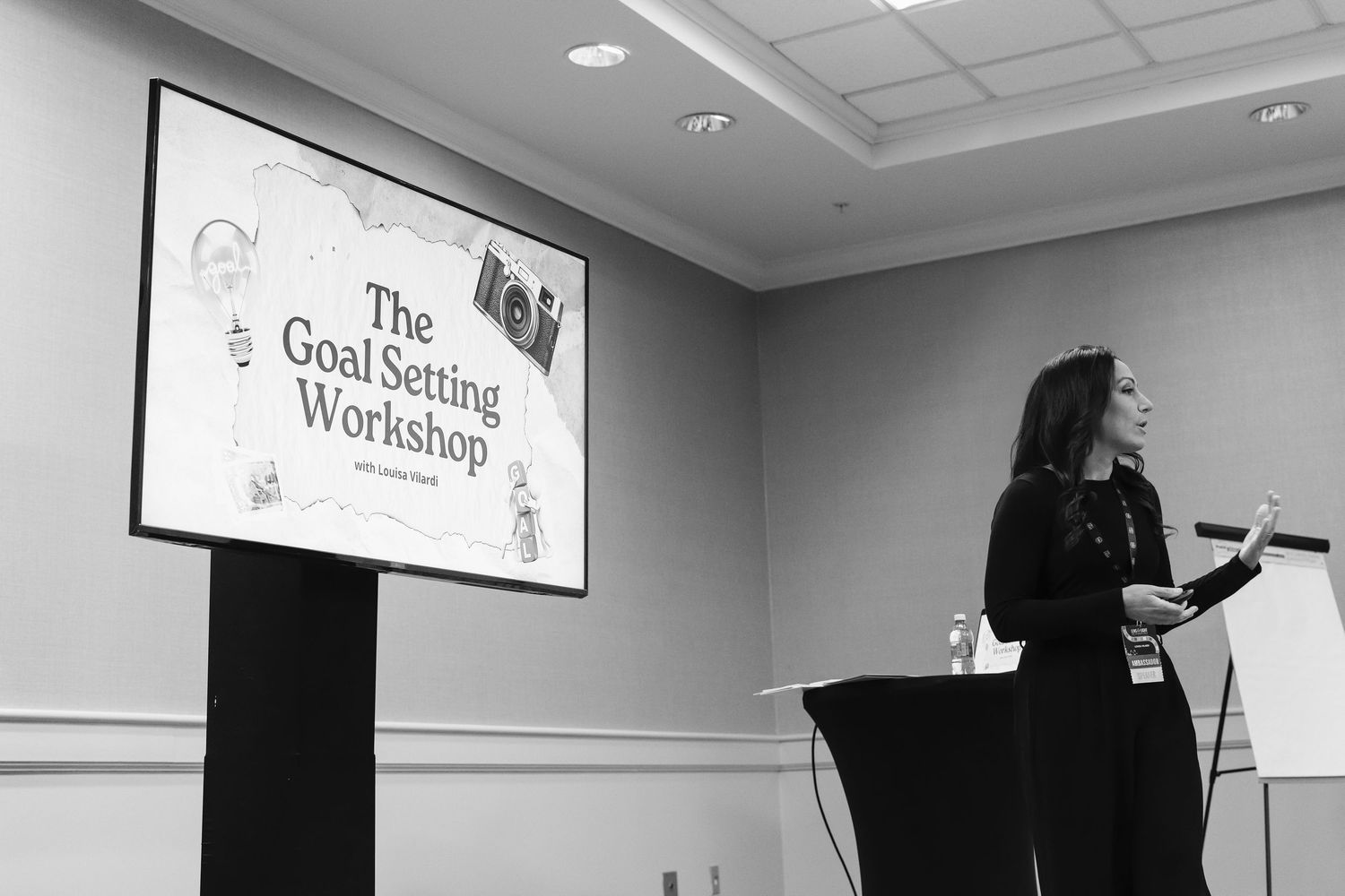 Woman presenting at The Goal Setting Workshop, standing at podium beside a large projected screen in a conference room.