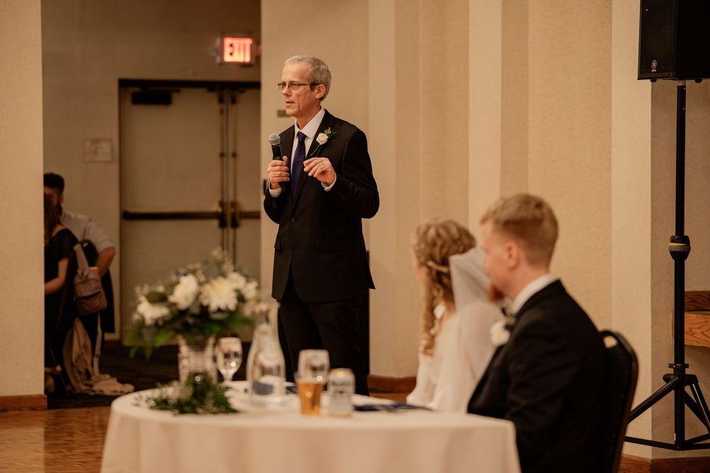 Man in suit giving a wedding toast speech at an elegant reception while bride and groom sit at the head table.