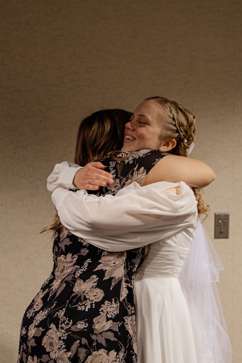 Bride in white dress and veil shares a joyful hug with a guest in a floral dress at a wedding.