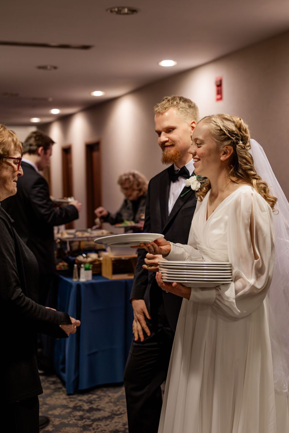 Bride in white gown holds plates while groom in black tuxedo smiles at wedding reception buffet.