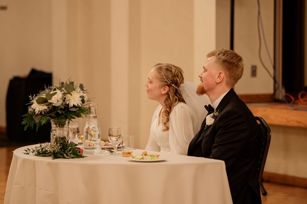 Bride and groom seated at sweetheart table with white floral centerpiece during elegant wedding reception dinner.