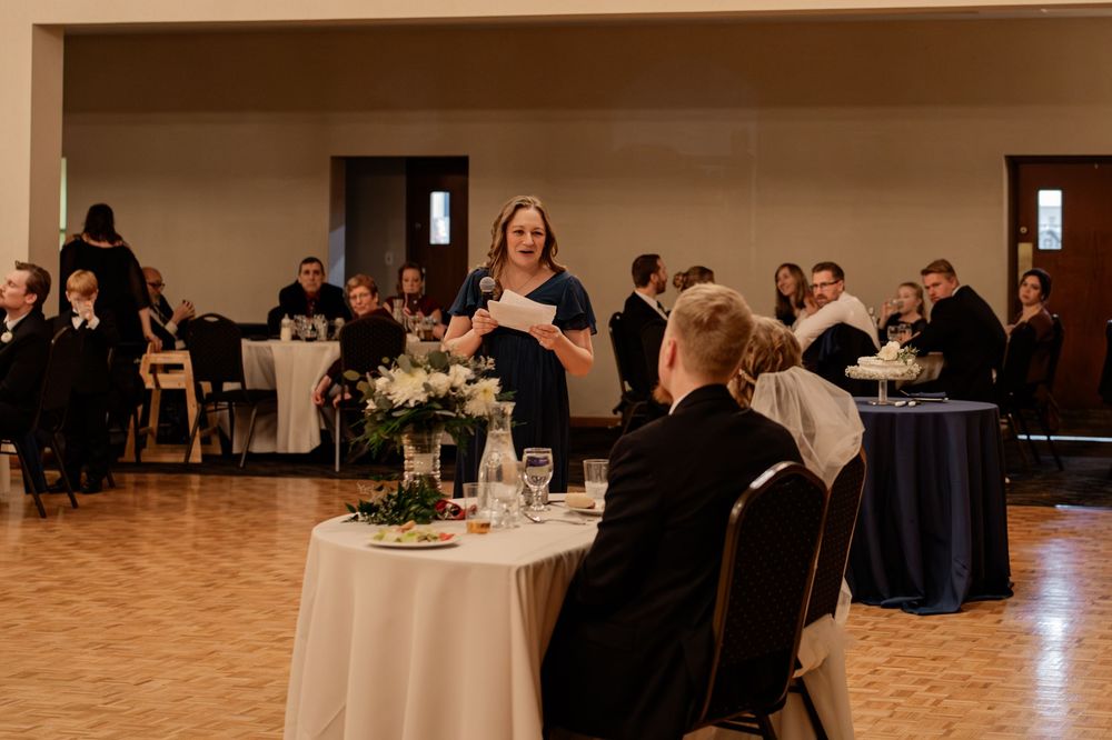 Woman in navy dress giving a speech at an elegant wedding reception with guests seated at white-clothed tables.