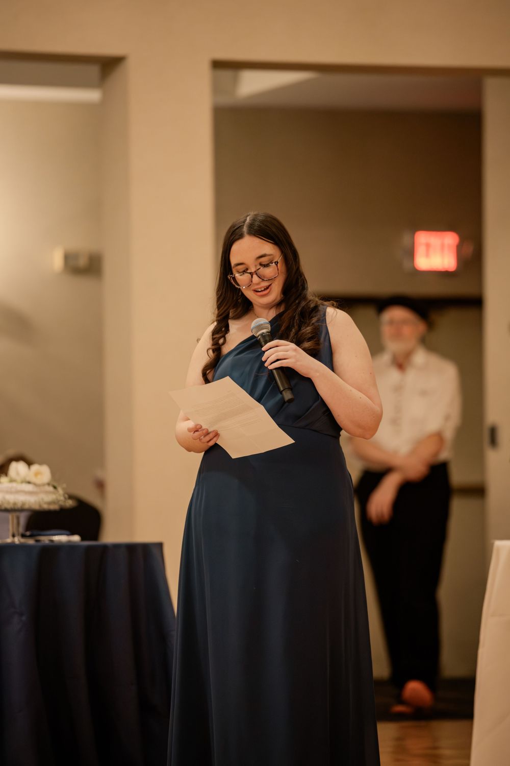 A woman in a navy gown gives a heartfelt speech at a wedding reception, holding a microphone and paper.