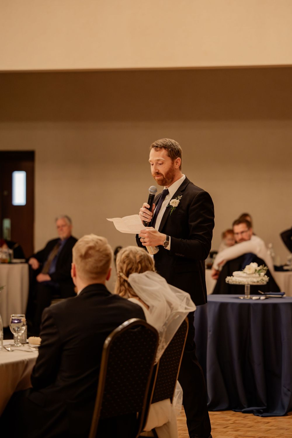 Man in suit giving a wedding speech at a reception, holding a microphone and paper while guests listen.