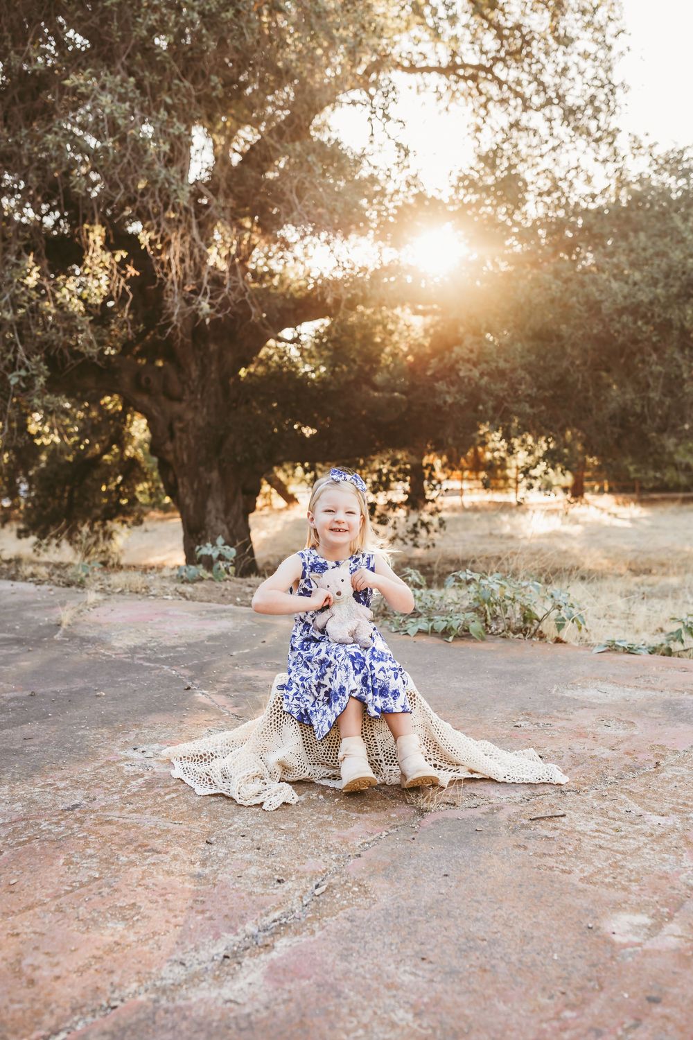 Toddler girl in blue floral dress holds stuffed animal on blanket outdoors with golden sunset light through oak trees.