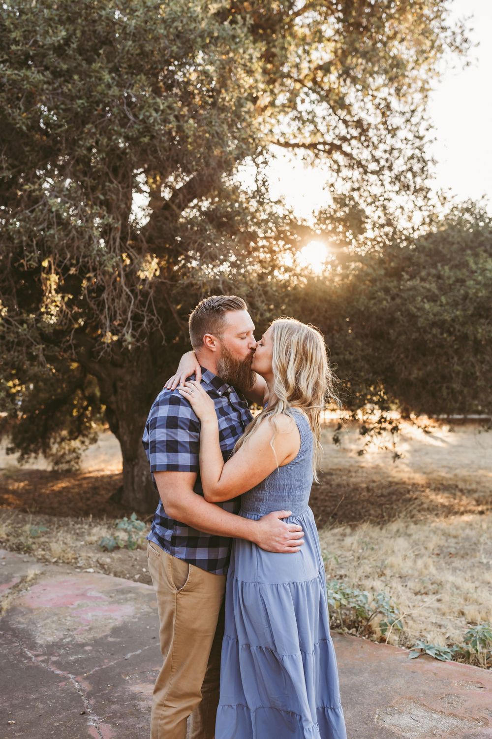 Couple sharing a romantic kiss at golden hour beneath oak trees, man in plaid shirt and woman in blue dress.