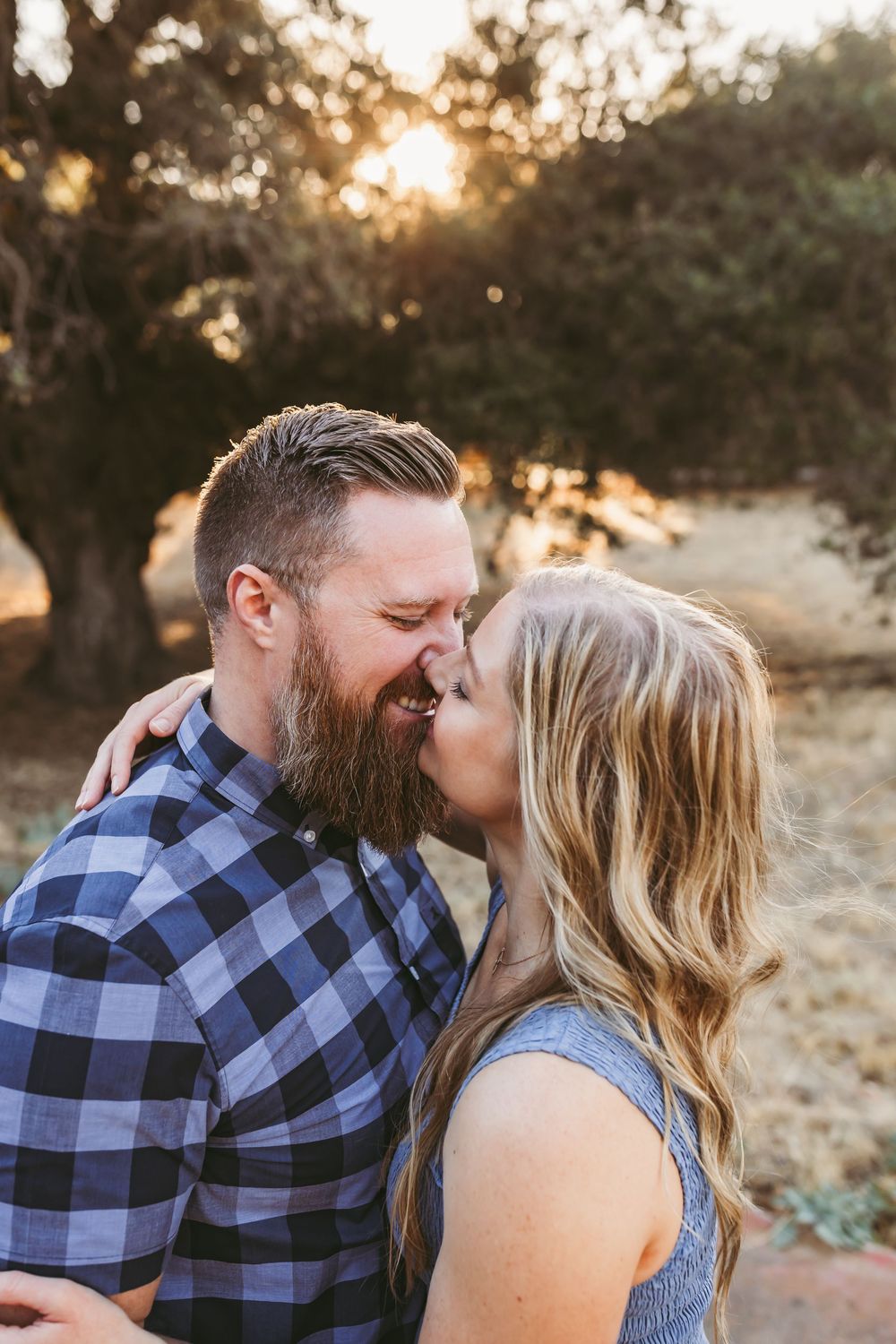 Couple sharing a romantic kiss outdoors at golden hour with trees and warm sunlight in the background.