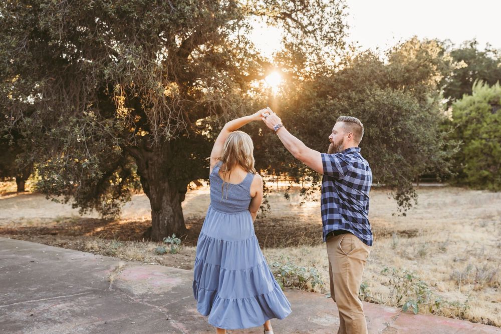 Couple dancing outdoors at golden hour, woman in blue dress being twirled by man in plaid shirt near trees.