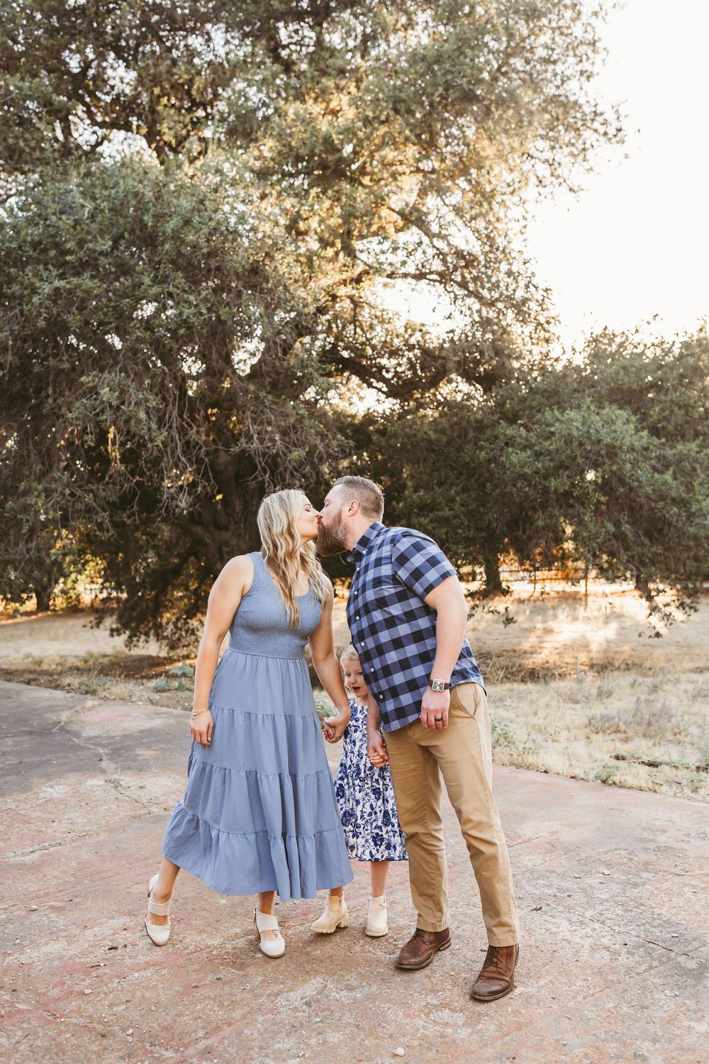 Couple sharing a kiss outdoors with their young daughter between them, surrounded by oak trees in golden light.