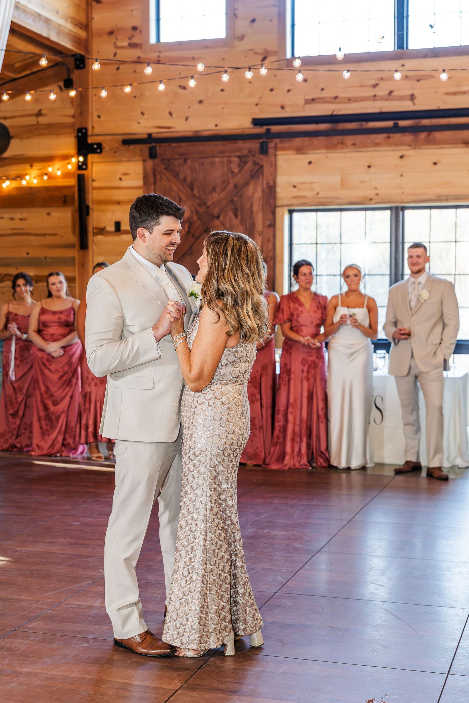 Bride and groom share first dance in rustic barn venue with bridesmaids in dusty rose gowns watching in background.