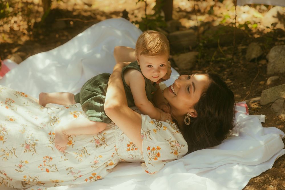 During a mini session photography, a mother lying on a white blanket outdoors, smiling while holding her happy baby in a green outfit.