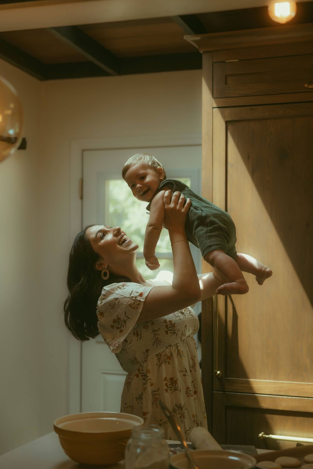 A joyful mother lifts her laughing toddler in a warmly lit rustic kitchen, sharing a playful moment.