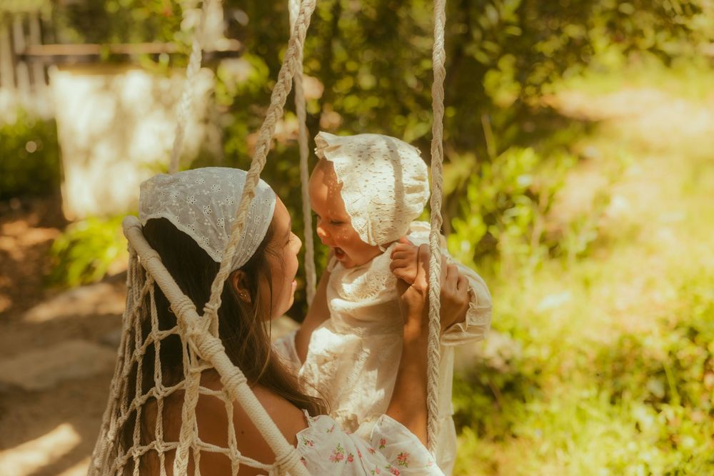 Mother in white lace holding baby in bonnet on rope swing outdoors in golden sunlight.