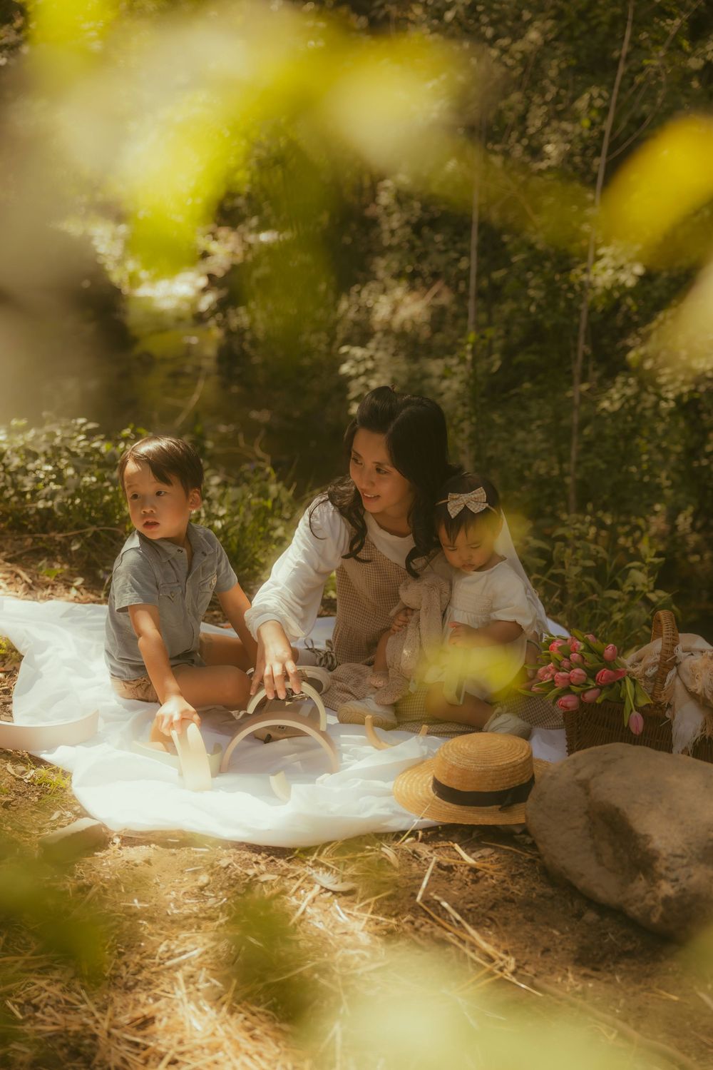 A mother and two young children enjoy a sunny outdoor picnic on a white blanket surrounded by lush greenery.