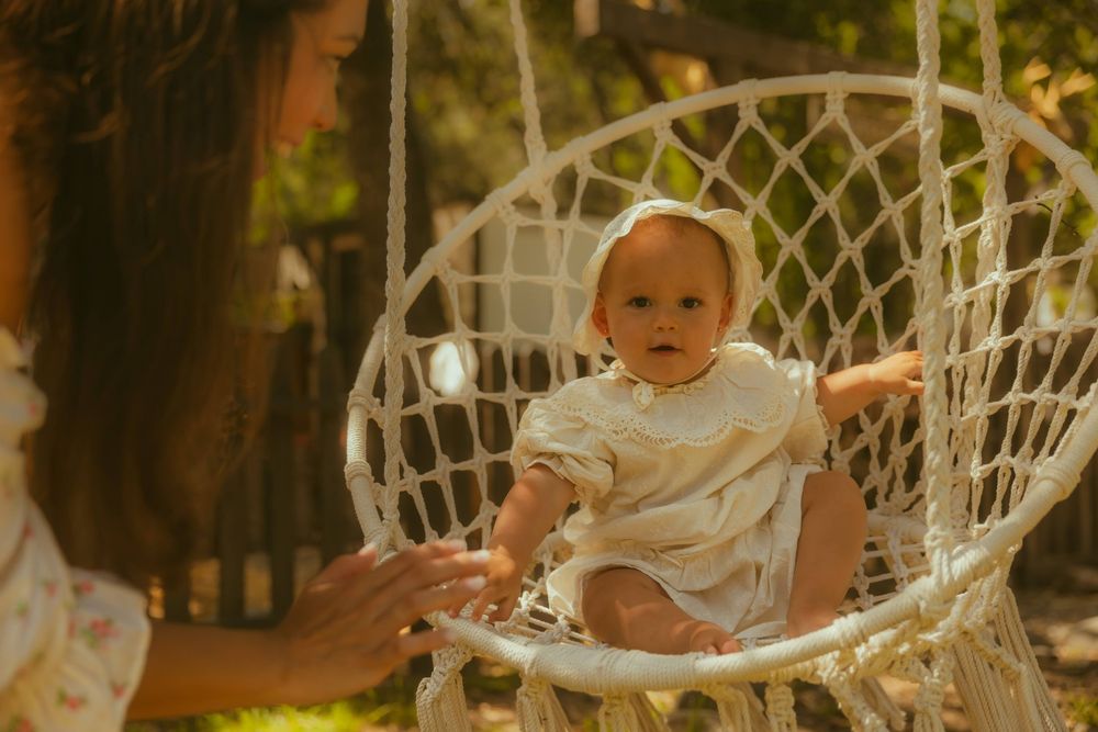 Baby sitting in a macrame hanging chair outdoors in golden sunlight, with a woman's hands reaching toward them.