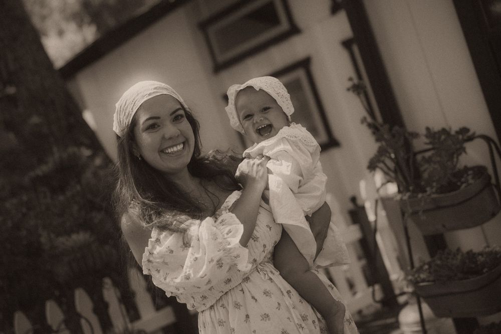 Smiling woman in white dress and headscarf holds a baby in bonnet outdoors in sepia-toned photo.
