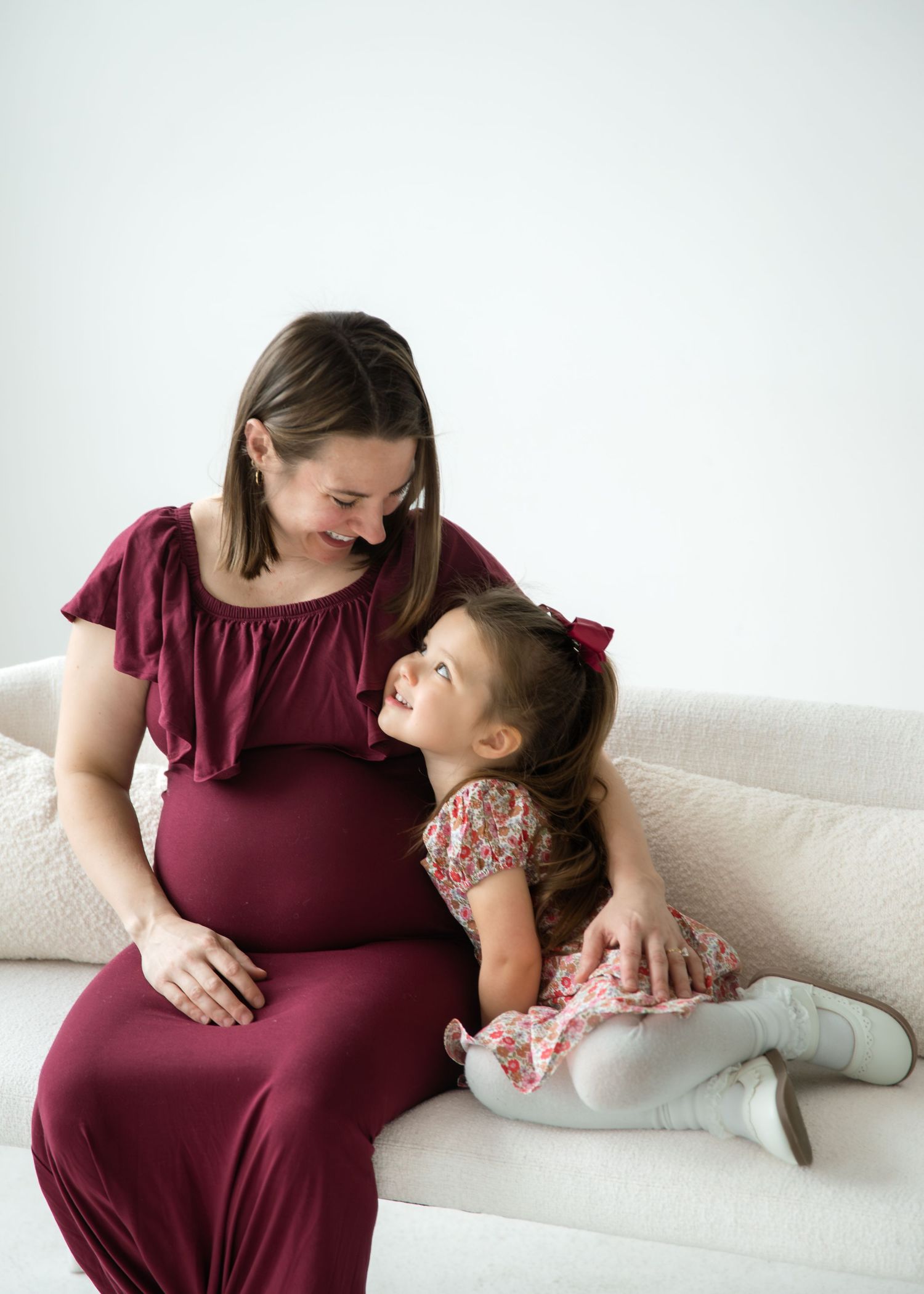 Pregnant woman in burgundy dress sitting on couch smiling at young girl in floral dress during maternity photo session.