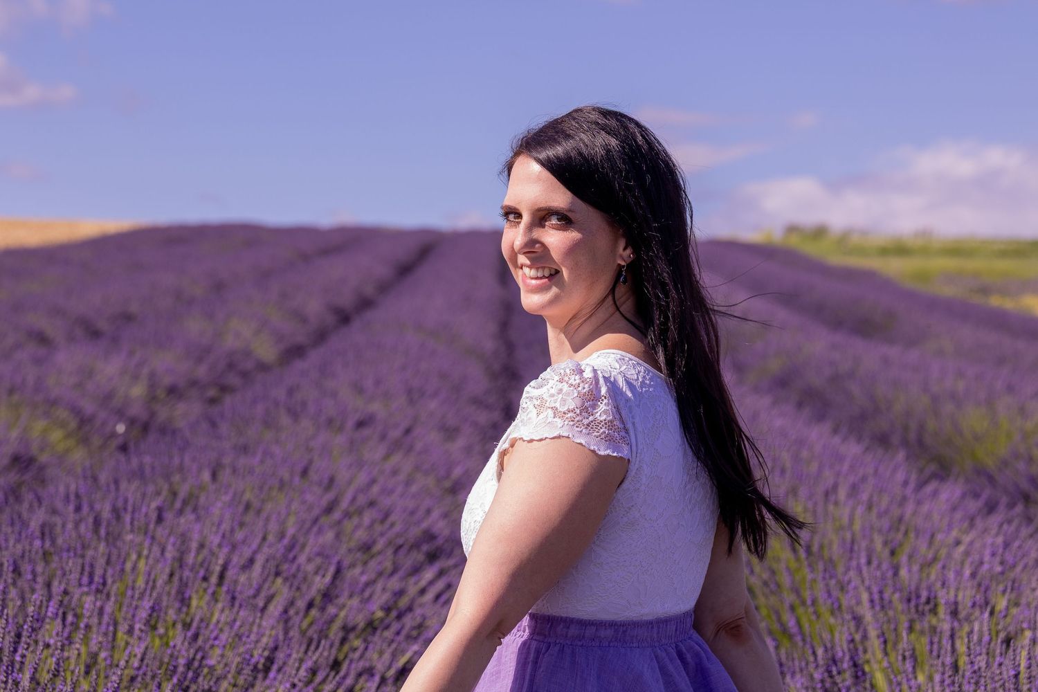 Smiling woman with long dark hair in white top and purple skirt standing in a vibrant lavender field under blue sky.