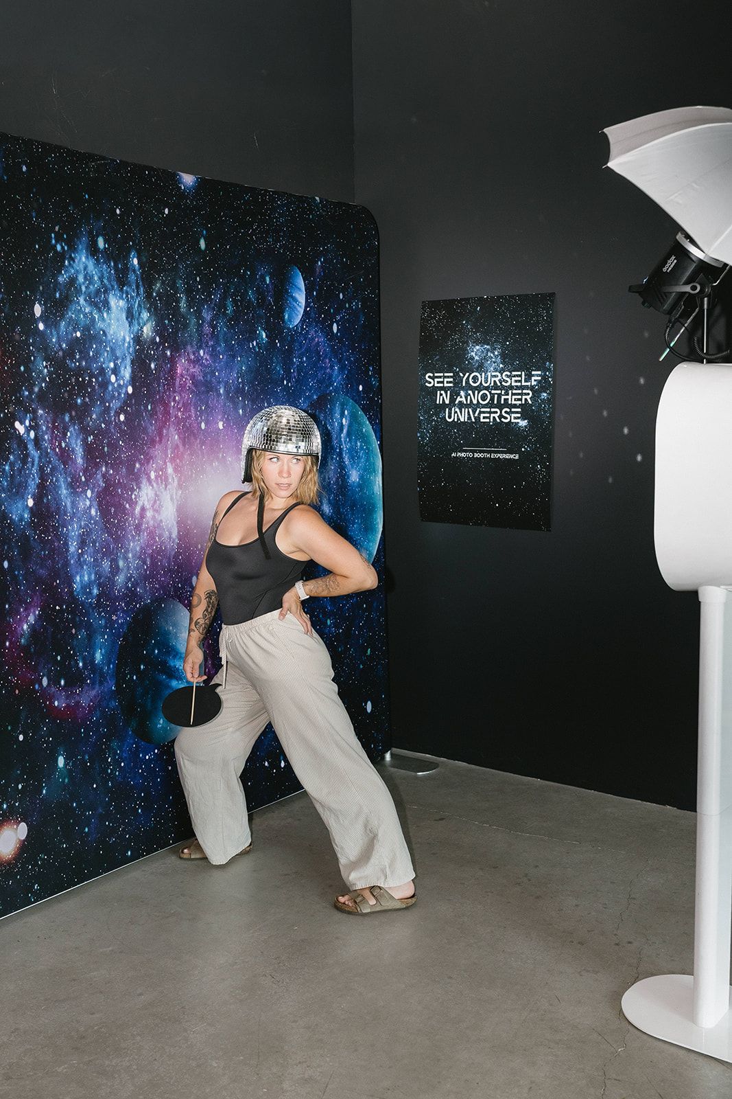 Woman posing playfully in front of a galaxy backdrop wearing a disco ball hat at a space-themed photo booth.
