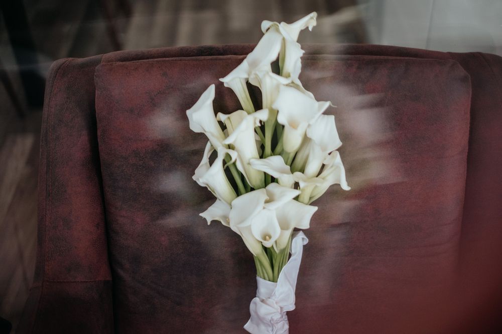 Elegant white calla lily wedding bouquet tied with ribbon resting on a dark red velvet chair.