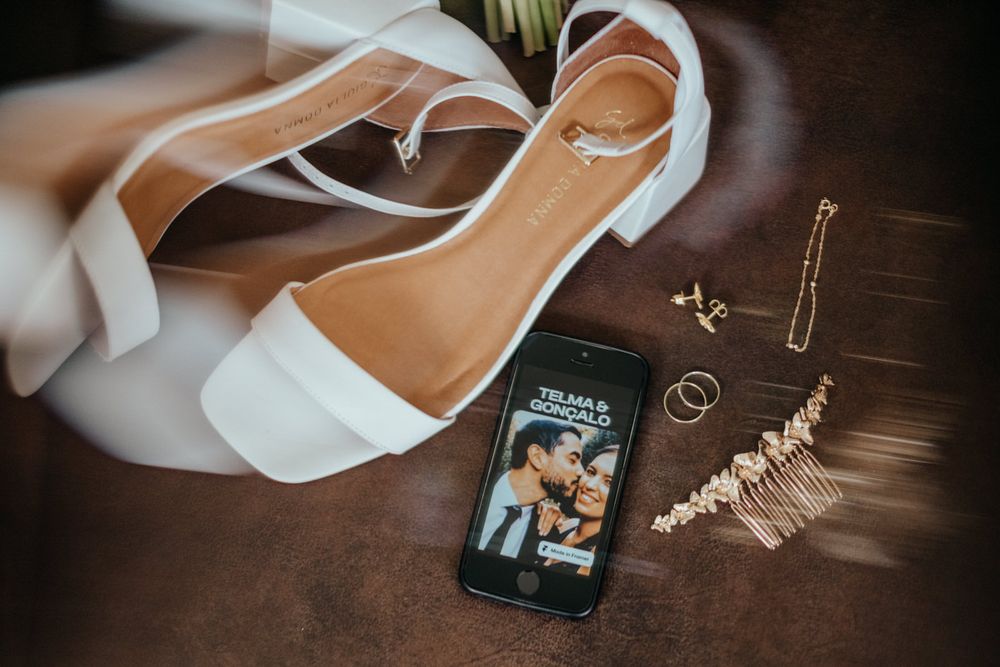 Flat lay of bridal accessories including white heels, gold jewelry, and a phone displaying wedding photos.