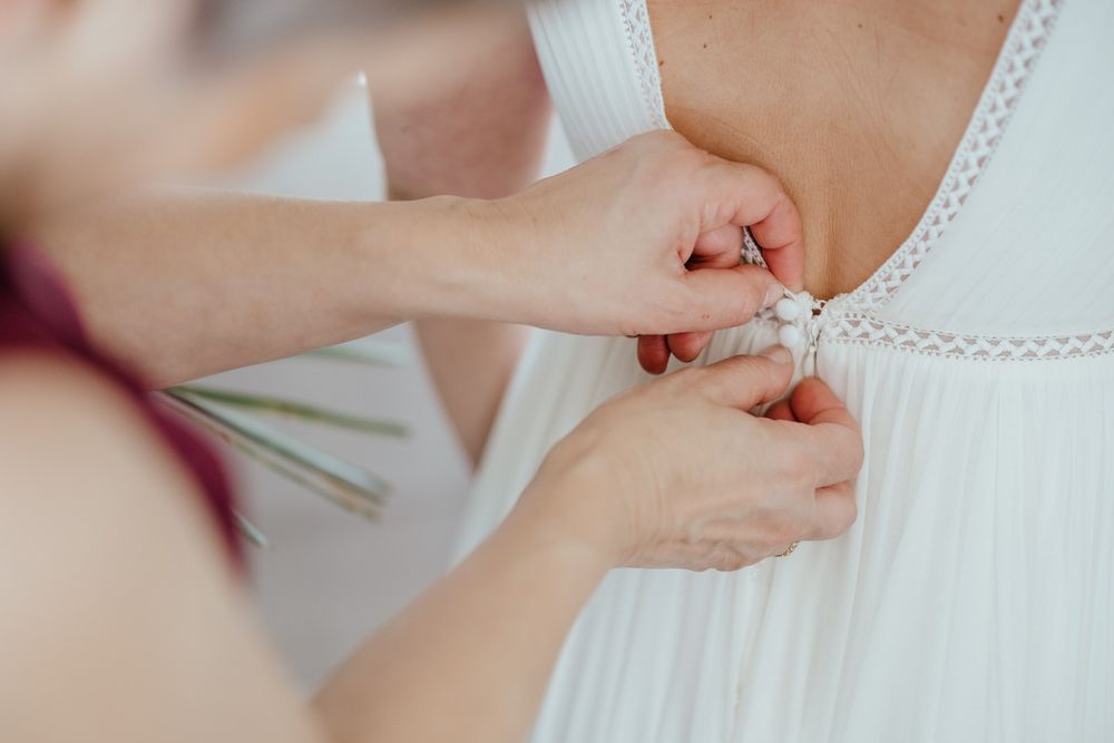 Close-up of hands buttoning the back of a white wedding dress during bridal preparations.
