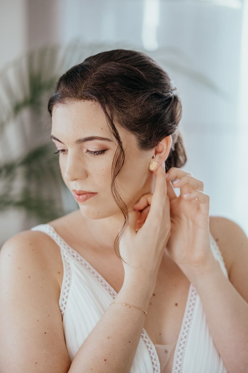 Bride in white dress adjusting gold stud earring, with elegant updo hairstyle and delicate bracelet.