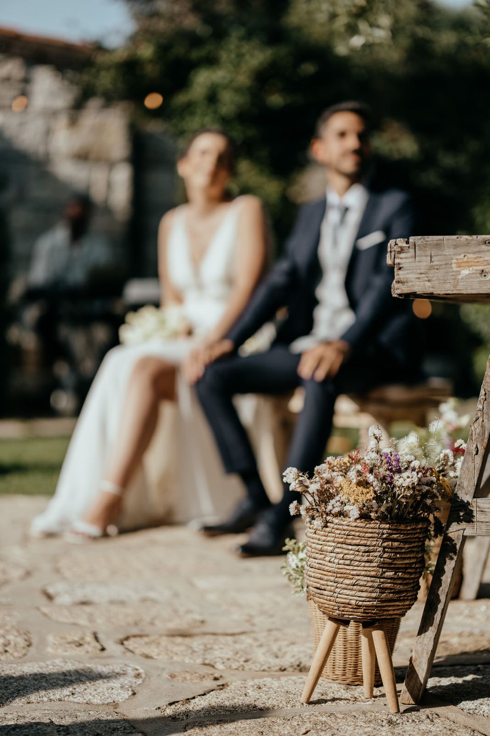 Bride and groom seated outdoors at rustic wedding ceremony with wildflower basket arrangement in foreground.