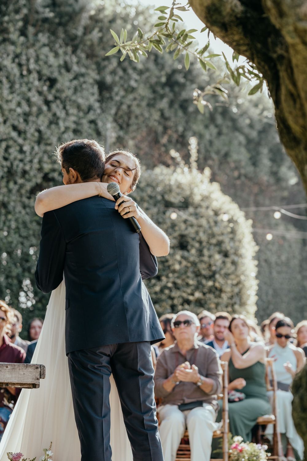Bride and groom embrace tenderly at outdoor wedding ceremony while guests applaud in background.