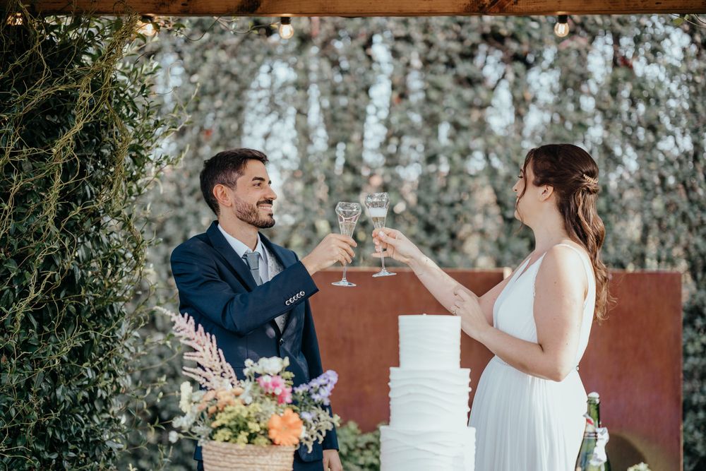 Bride and groom toasting with champagne glasses near white wedding cake at outdoor garden reception.