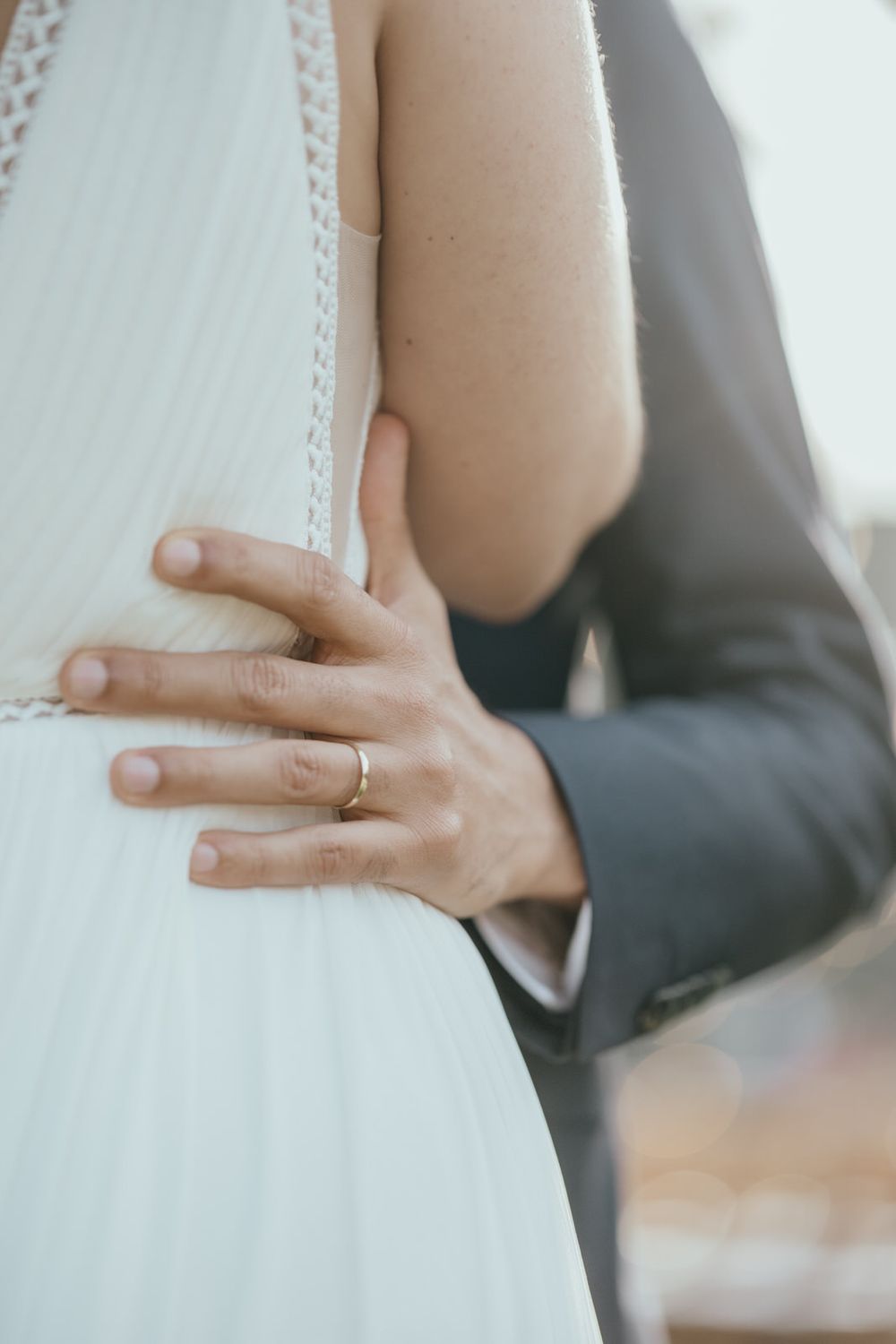 Groom in gray suit placing hand on brides white pleated wedding dress waist, wearing a gold wedding band.