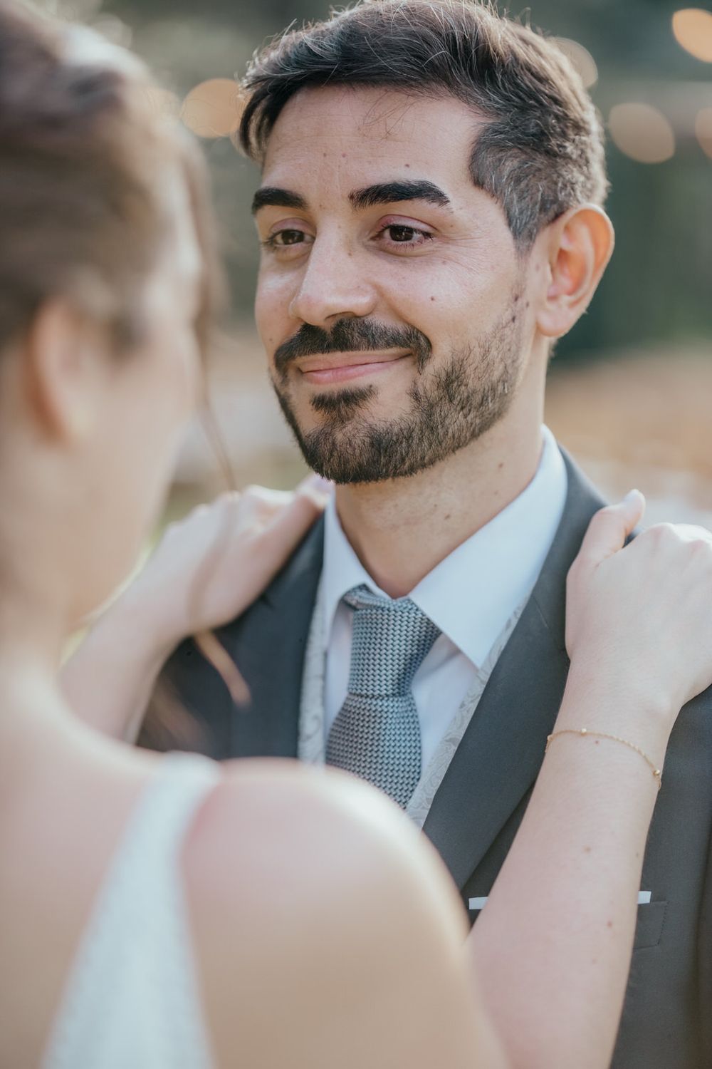 Smiling bearded groom in gray suit and tie as bride adjusts his collar during wedding ceremony.