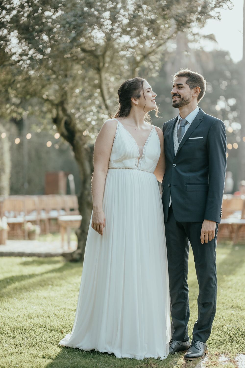 A bride in a white flowing gown and groom in a navy suit share a romantic moment outdoors at a wedding reception.
