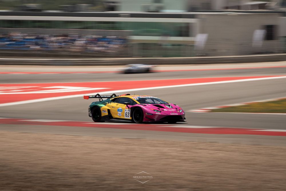 Pink and yellow race car speeding on a circuit track during a motorsport event on a sunny day.